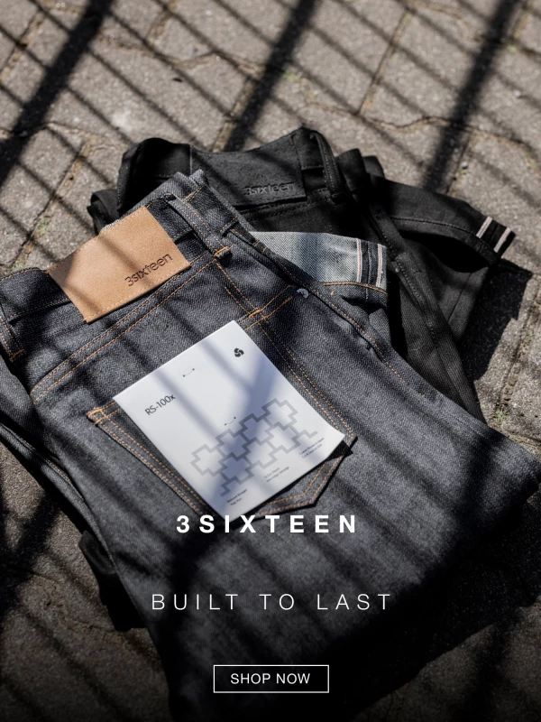 Close-up of folded 3sixteen denim jeans with a tan leather patch on the waistband, placed outdoors on cobblestone. Text reads 3SIXTEEN BUILT TO LAST and a SHOP NOW button appears in the corner. 