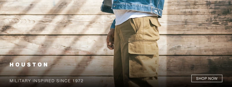 A person wearing khaki cargo pants, a white shirt, and a denim jacket stands in front of a wooden wall. Text reads HOUSTON and MILITARY INSPIRED SINCE 1972. A SHOP NOW button appears on the right. 