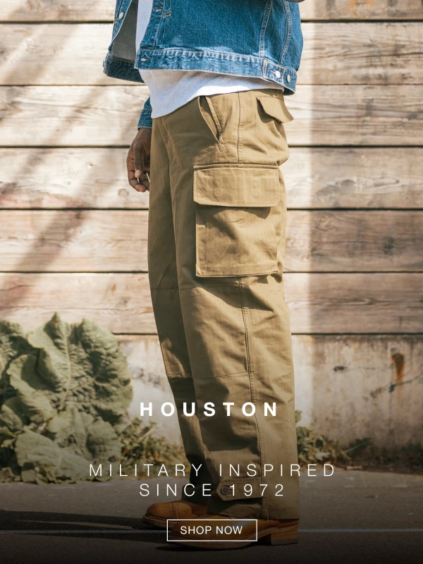 A person wearing khaki cargo pants, a white shirt, and a denim jacket stands in front of a wooden wall. Text reads HOUSTON and MILITARY INSPIRED SINCE 1972. A SHOP NOW button appears on the right. 