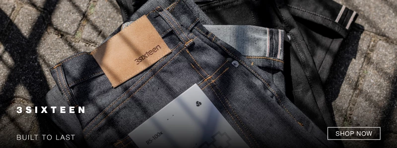 Close-up of folded 3sixteen denim jeans with a tan leather patch on the waistband, placed outdoors on cobblestone. Text reads 3SIXTEEN BUILT TO LAST and a SHOP NOW button appears in the corner. 